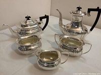 Full view of five-piece silver plate tea set including teapot, coffee pot, creamer, sugar bowl, and waste bowl with black handles.