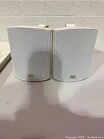 Pair of white Enforcer brand indoor/outdoor speakers with white metal mesh grilles, shown front-facing on a table against a wall.