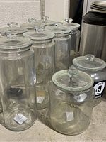 Front and angled view of 8 tall and 2 short clear glass jars with lids, showing their cylindrical shape and glass knob tops.