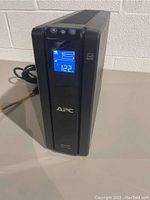 Front angled view of APC Back-UPS Pro 1500 UPS unit showing LCD display active with voltage and battery icon, power cable coiled at back.
