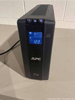 Front and angled view of APC Back-UPS XS 1000 UPS unit powered on showing the LCD display with input voltage 122V and battery status.
