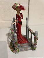 Front angle of figurine showing woman in red dress, floral hat, holding Coca Cola glass, standing on stone platform with railing wrapped in pink roses.