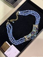 Full view of the triple-strand blue faceted crystal bead necklace with a multi-gemstone and rhinestone decorated gold-tone centerpiece inside a black velvet-lined box.