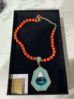 Heidi Daus necklace displayed in a black box showing bright orange round beads and a geometric pendant with blue faceted stone and turquoise enamel, with certificate.