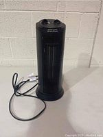 Black ceramic tower heater standing against a white wall, showing front and side view with power cord coiled on the floor.