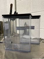 Front and side angle of two clear rectangular plastic storage containers with black lids, showing transparent sides and black lids.