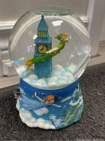 Snow globe showing Peter Pan flying in front of a clock tower inside glass globe, sitting on a detailed painted base with Peter Pan characters and clouds scene