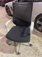 Front view showing black mesh backrest, black fabric seat, white armrests, and caster wheels on concrete floor.
