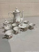 Photo of full tea set including teapot, sugar bowl, six teacups and six saucers on a gray surface with a beige wall background.