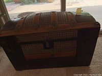 Front view of the dome top steamer trunk showing the rounded woven texture surface, wood slats, and metal accents on a dark colored body.