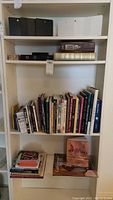 Shelving unit containing a variety of over 30 books standing upright and stacked, alongside a set of four black metal bookends on a separate shelf.
