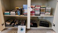 Wide shot of white cabinet with shelves holding a majority of the cassette tapes. Visible cassettes on shelves, some stacked, some lined up with labels visible. Includes a few boxed sets such as The Four Gospels and Amos N Andy tape collections.