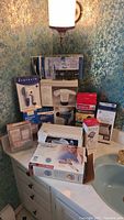 Photo showing assortment of boxed home health items arranged on bathroom counter including Waterpik, heating pads, hair dryer, blood pressure cuff and more.