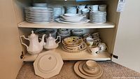 Shelving unit with multiple stacks of assorted china including plates, cups, saucers, and teapots in cream color and some colorful floral patterned items.