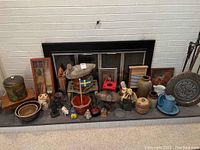 Wide view of assorted decorative pottery, figurines, fireplace tools, and framed artwork arranged on a fireplace hearth