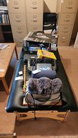 Full view of the folding massage table with various accessories and boxes on top, showing black cushioned surface and wooden frame.