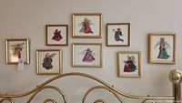 Photo showing the full set of eight framed needlepoint angel artworks arranged on a wall above a bed frame, light gold frames visible with varied angel figures in needlepoint textile art.