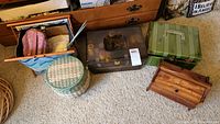 Overview photo showing several sewing baskets and accessories on carpeted floor including blue fabric bag, metal chest, green hard case, round woven basket, and wooden box with drawers.
