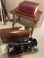 Overview showing sewing machine mounted on wooden base with power cord, wooden carrying case open in front, instruction manual, end table and two reference books.