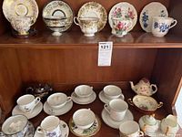 Wide view of thirteen tea cups with matching saucers arranged on two shelves and two teapots visible.