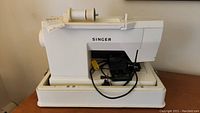 Full side view of vintage Singer sewing machine in white plastic carrying base with spool pin and power cord.