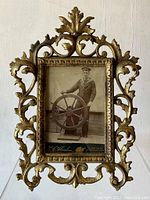 Front view of ornate cast metal frame holding black-and-white photograph of a sailor at a ship’s wheel.