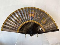 Front view of an antique painted hand fan with dark background and gold, blue, and pink floral designs.