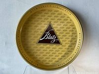 Top-down view of the full circular Blatz beer tray showing the central brown triangle logo and the repeating Blatz script pattern on a gold background.