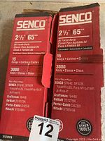 Two red and white boxes labeled Senco showing model DA25EPB, 15 gauge, 2 1/2 inch (65mm) finish nails, each box with 3000 nails. Boxes list compatible power tool models.