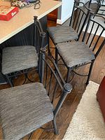 Four black metal frame bar height stools with gray fabric cushions positioned on wooden floor near kitchen island.