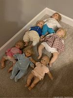 Photo showing six dolls ranging 15 to 19 inches in various clothing and hair styles lying on carpeted floor.