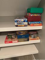 Shelf showing stacked board games and puzzle boxes including Life, Sorry, Battleship, Connect Four, Hangman, Monopoly, a 1000-piece puzzle, and decorative tins