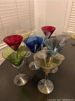 Nine colored martini glasses arranged on a dark surface showing their wide bowl and stem design.