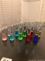 Front view of 11 assorted colored shot glasses with Royal Caribbean logos and one without logo, on black table.