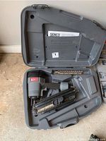 Senco pneumatic nailer in gray and black inside an open plastic carrying case, with coil of nails and instruction manual.