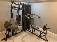 Full view of the gray and black Powerhouse multi-station weight machine against a beige wall in a carpeted room showing multiple workout handles, padded seats, and weight stack.