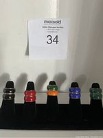 Front view photo showing six enamel rings mounted on black velvet ring display stand in black, red, green, orange, blue colors.
