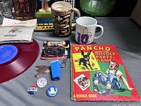 The photo shows assortment of vintage collectibles including a red vinyl record album, a 'Pancho Pin the Donkey' party game book, two ceramic mugs (tiki style and white with number 10), Matchbox car set in original packaging, some metal and plastic keychain items, and buttons.
