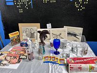 Photo of assorted collectibles and memorabilia on table including wooden dolphin statue, unopened trading card packs, glassware, prints, and boxed craft kit.