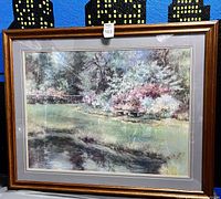 Framed landscape print featuring a peaceful river scene with trees and flowers in soft pastel colors.