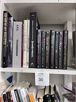 Books shelved tightly on a white shelf, mixed hardcover and paperback, visible titles including 'Transmission', 'Country Look', 'The Art of the Fold', and multiple black hardcover books in a fantasy series.