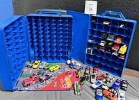 Two blue Hot Wheels carrying cases opened, displaying the multiple compartments designed for Hot Wheels toy cars. Several cars are played out in front including various models and colors, and a red Hot Wheels branded cloth racing mat is laid out.