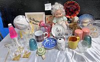 Full lot view showing various items on table including glassware, plates, dolls, and plush toys against black backdrop