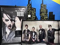 Two framed music posters hung against a blue and black painted wall with cityscape design, Marilyn Manson on left, AFI Crash Love on right