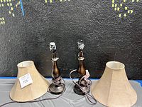 Two wooden lamp bases and two beige fabric shades placed on a table against a black wall with city night lights art.