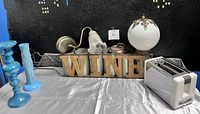 Photo showing the metal wine rack with wooden letters spelling 'WINE' along with two blue glass candlesticks, a silver toaster, a lamp with a frosted glass shade, and a lamp with a white globe shade and brass accents.