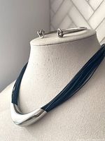 Necklace displayed on a mannequin showcasing multiple strands of midnight blue leather with a central large curved silver tone tube pendant and matching silver tone knot bangle above.