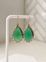 Front view of two pear-shaped faceted green onyx stones with vermeil gold decorative borders attached to gold ear hooks, displayed on white earring stand.