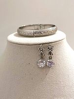 Vintage silver tone hinged floral etched bracelet displayed on a stand with rhinestone dangle earrings