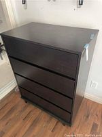 Full view of black 4 drawer dresser with no handles, standing on wooden floor beneath light wall.